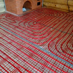 Sergii Cholavyn - Czerwone rury ogrzewania podłogowego marki Rommer, ułożone na siatce w pomieszczeniu z drewnianymi ścianami i ceglanym kominkiem, gotowe do zalania wylewką.