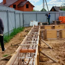 Zbrojenie fundamentów w drewnianym szalunku, widoczne rury kanalizacyjne wewnątrz konstrukcji, w tle dom z czerwonej cegły i pracownik budowlany przy ogrodzeniu.