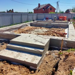 Sergii Cholavyn - Wykop pod fundamenty domu jednorodzinnego z widocznymi schodami wejściowymi, częściowo zalany betonem, z pracownikami w tle, na tle ogrodzenia i budynku w stanie surowym.