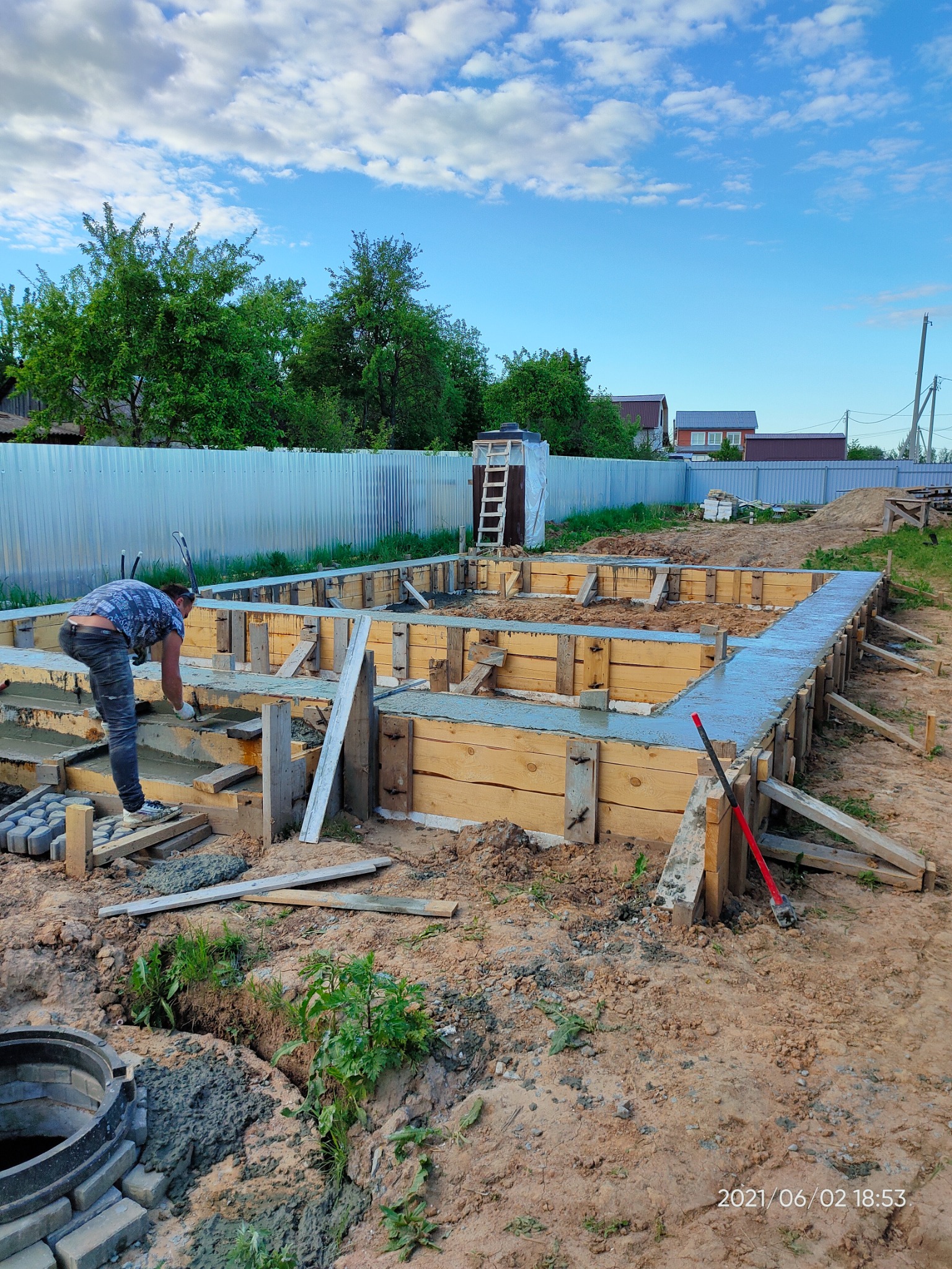 Wylewanie betonowej ławy fundamentowej w drewnianym szalunku. Widoczny pracownik wyrównujący powierzchnię betonu, czerwona łopata oraz studzienka kanalizacyjna w wykopie.