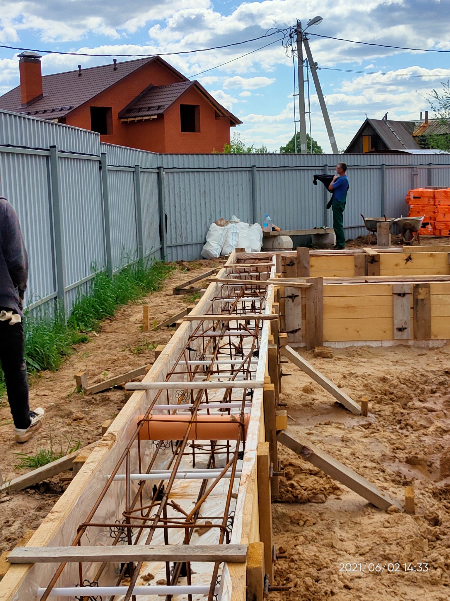 Zbrojenie fundamentów w drewnianym szalunku, widoczne rury kanalizacyjne wewnątrz konstrukcji, w tle dom z czerwonej cegły i pracownik budowlany przy ogrodzeniu.