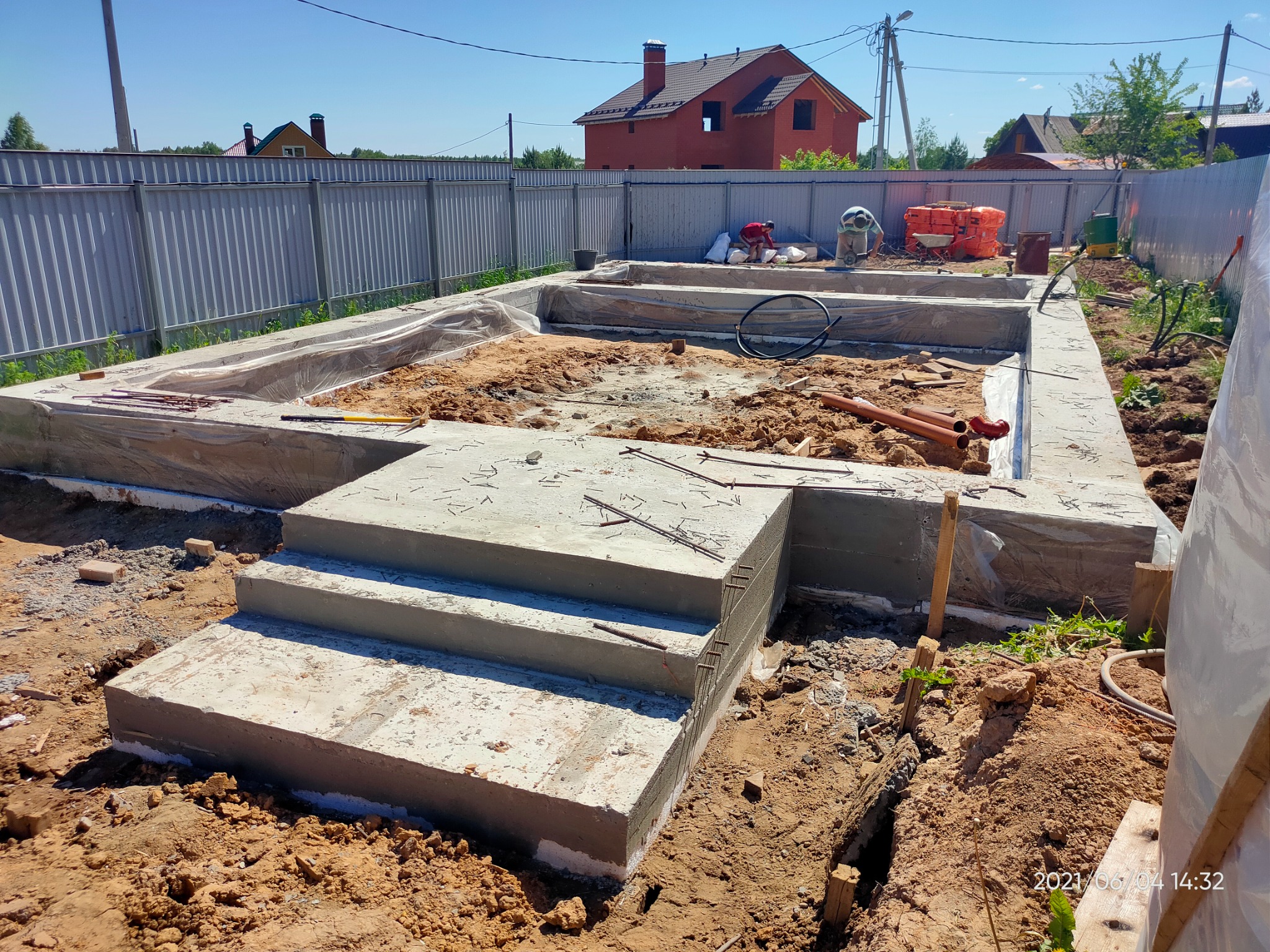 Wykop pod fundamenty domu jednorodzinnego z widocznymi schodami wejściowymi, częściowo zalany betonem, z pracownikami w tle, na tle ogrodzenia i budynku w stanie surowym.