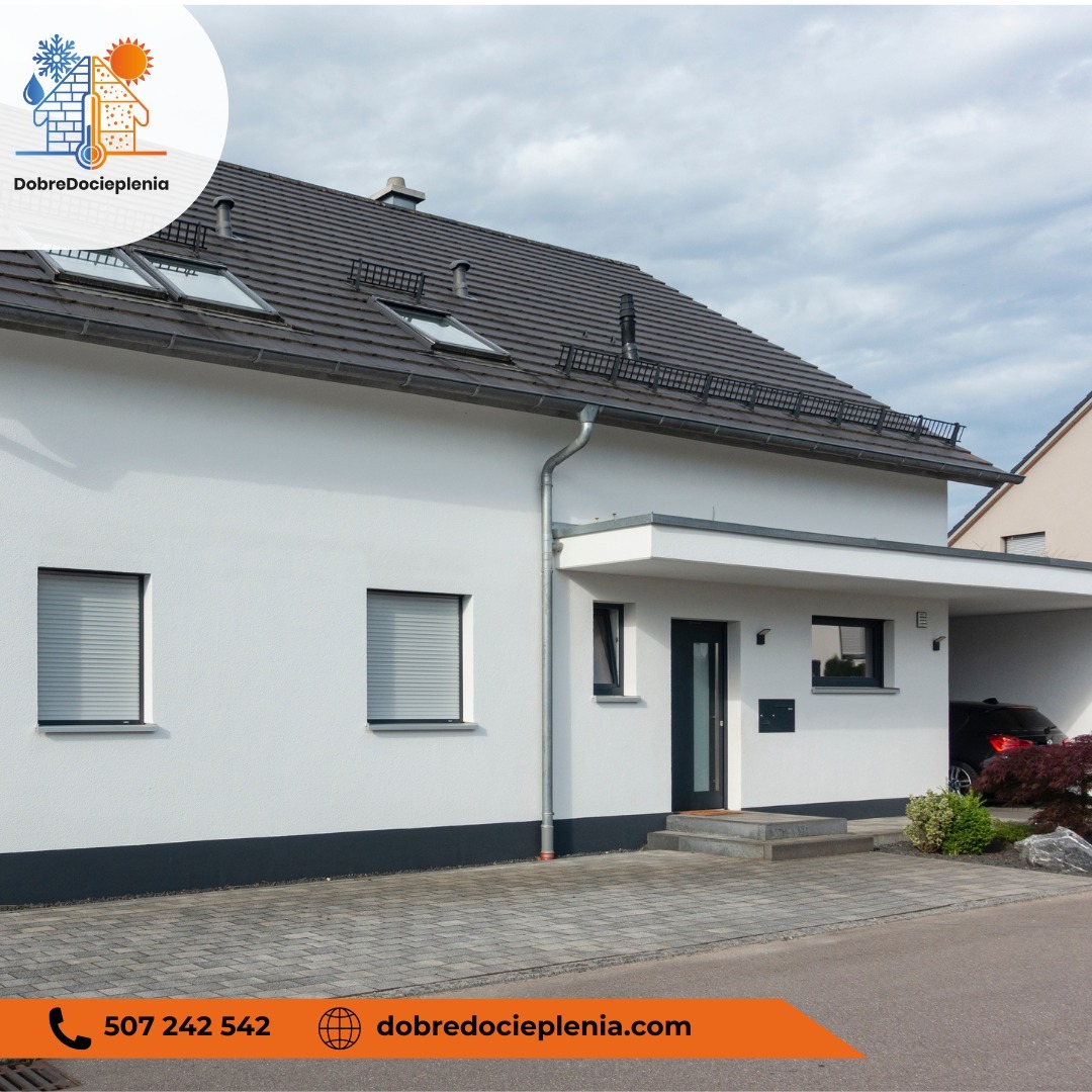 Biały dom jednorodzinny z szarą dachówką i roletami zewnętrznymi w oknach, widok z poziomu chodnika z kostki brukowej. Budynek z podjazdem i zadaszeniem dla samochodu.