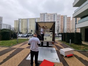 Załadunek mebli i kartonów do samochodu dostawczego na tle bloków mieszkalnych. Pracownicy w białych koszulkach w trakcie przenoszenia wyposażenia.