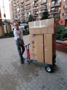 Mężczyzna w okularach i białej koszulce przewozi wózkiem skrzynie kartonowe na tle bloku mieszkalnego, kostka brukowa na chodniku.