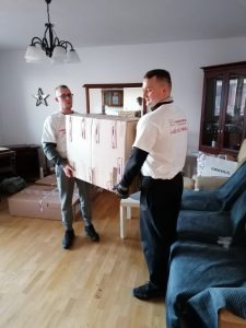Dwóch mężczyzn w białych koszulkach z logo firmy transportowej niesie duże, zaklejone kartonowe pudło w jasnym pokoju z parkietem.