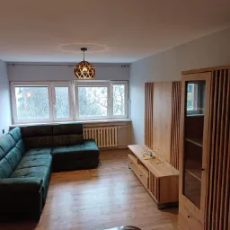 Nowoczesny salon z zielonym narożnikiem, drewnianą zabudową meblową z lamelami i geometryczną lampą wiszącą.