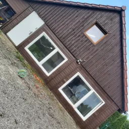 Dom z brązową drewnianą elewacją, białymi oknami i drzwiami garażowymi, widziany pod kątem z ziemi, z widocznym zwiniętym kablem elektrycznym i częścią ogrodzenia.