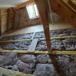 Izolacja poddasza wełną mineralną między krokwiami, widoczne folia paroizolacyjna i deski podłogowe, okno dachowe w tle.