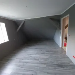 Szare, puste poddasze z drewnianą podłogą, skośnymi ścianami i widocznymi elementami konstrukcyjnymi, przygotowane do dalszego wykończenia.