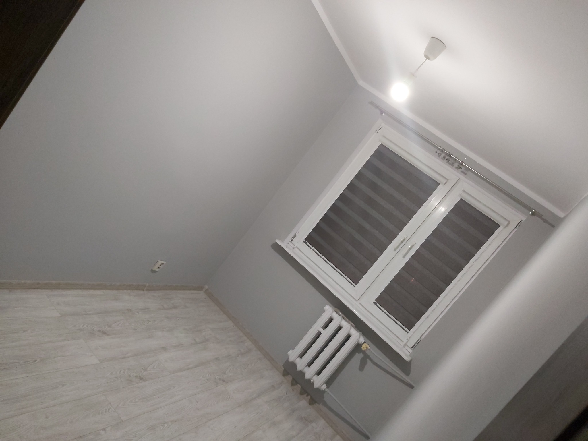 Szare wnętrze pokoju po remoncie z panelami podłogowymi, białym oknem z żaluzjami, grzejnikiem pod oknem i białym sufitem z lampą.