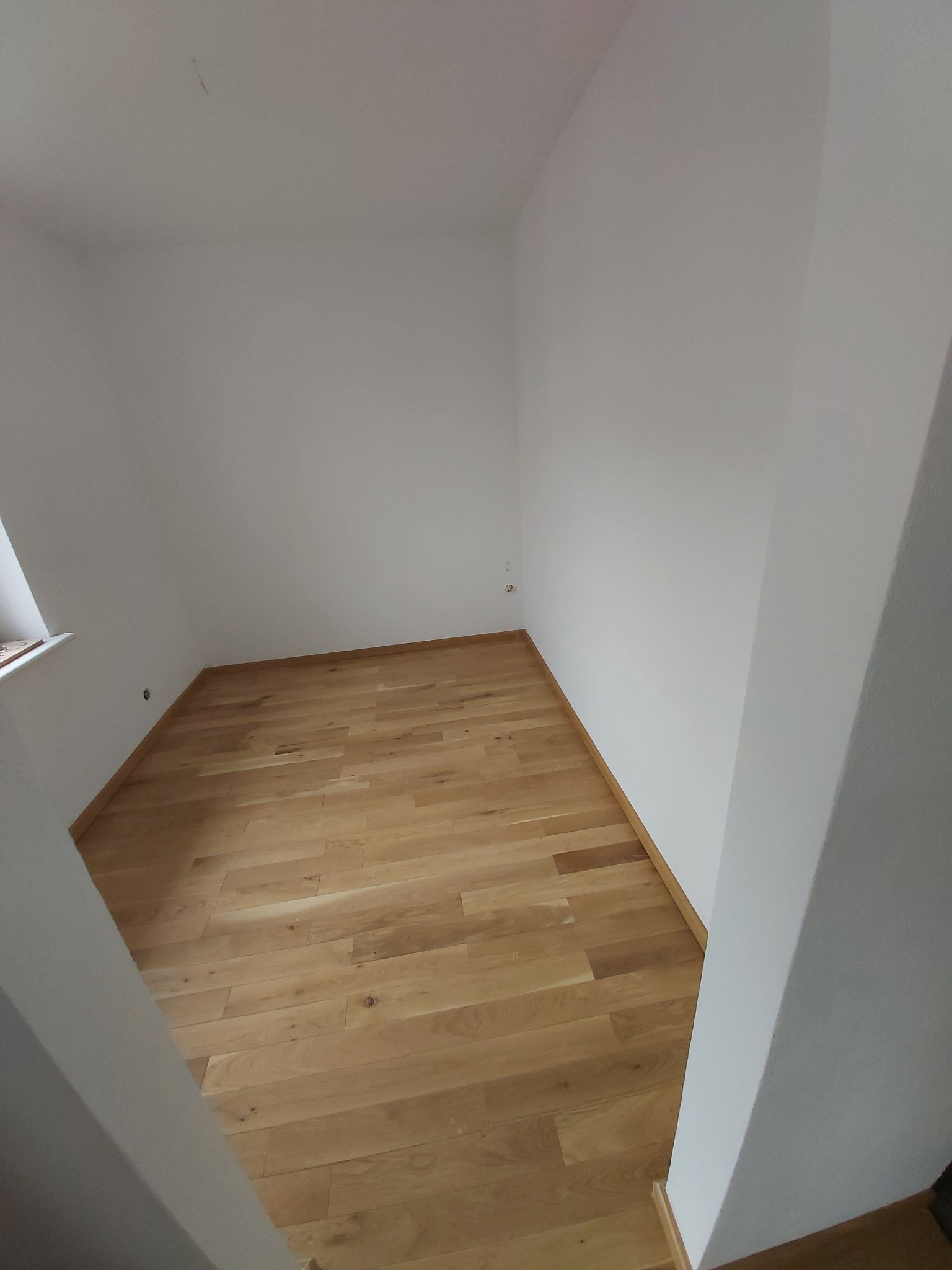 Puste pomieszczenie z parkietem ułożonym w jodełkę, świeżo pomalowane na biało ściany, widoczny fragment okna po lewej stronie.