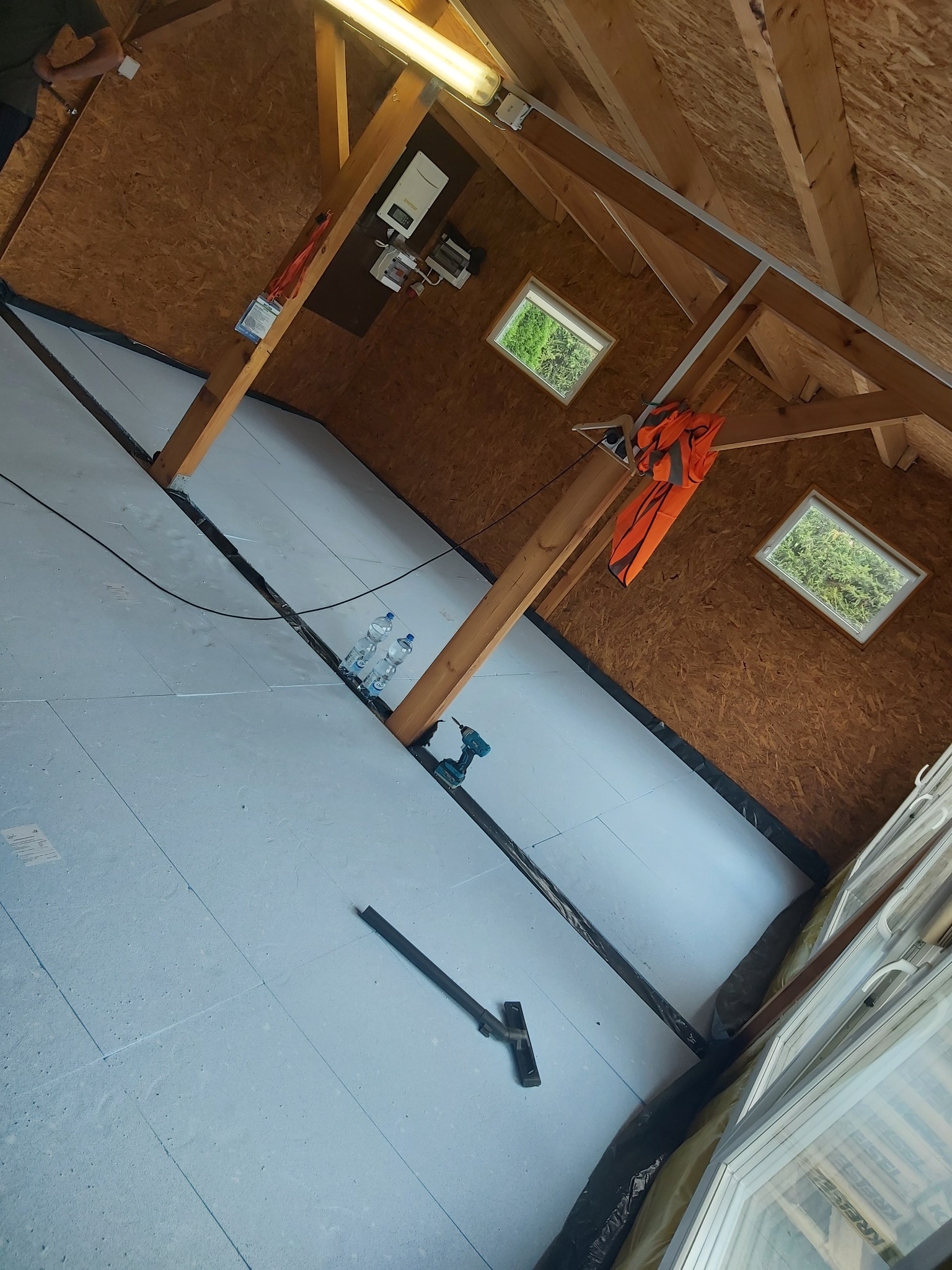 Poddasze z widoczną izolacją termiczną, narzędziem do docinania i wiertarką akumulatorową, butelki z wodą w tle, widok z dołu na drewnianą konstrukcję dachu i okna.