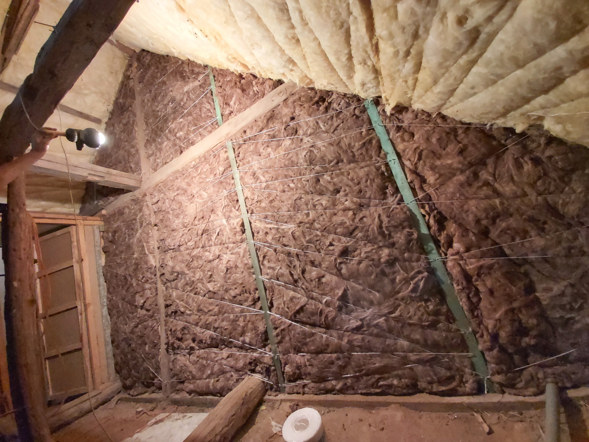 Ocieplenie poddasza wełną mineralną, zabezpieczone sznurkiem i zielonymi listwami, widoczny fragment drewnianej konstrukcji dachu i oświetlenie robocze.