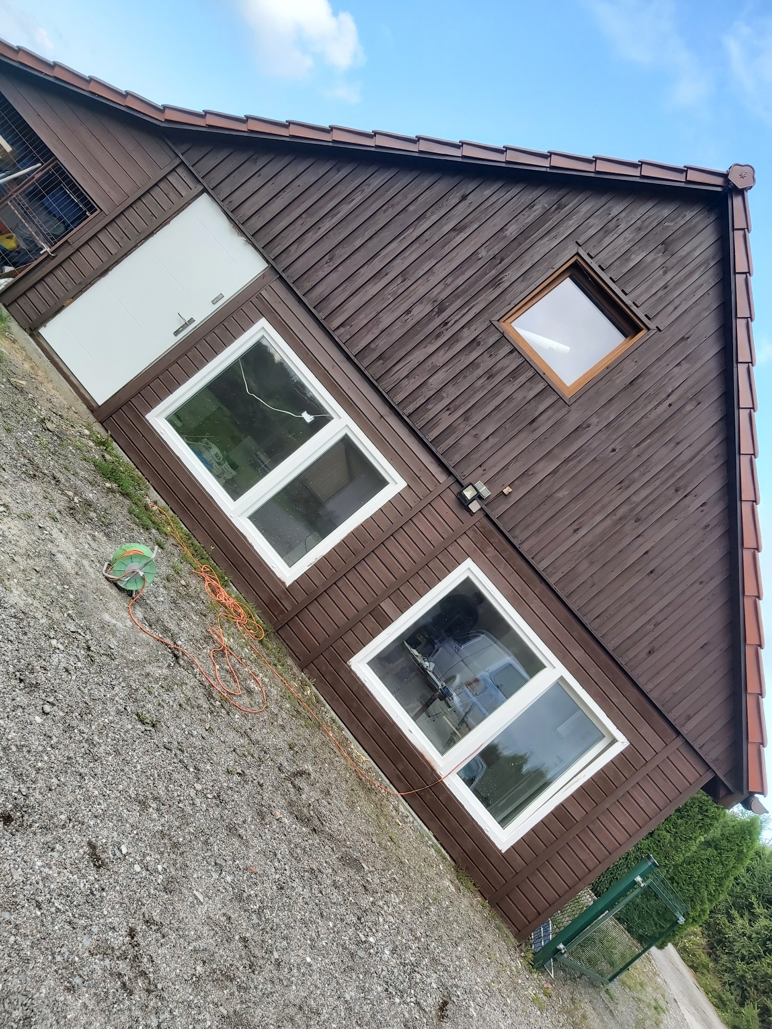 Dom z brązową drewnianą elewacją, białymi oknami i drzwiami garażowymi, widziany pod kątem z ziemi, z widocznym zwiniętym kablem elektrycznym i częścią ogrodzenia.