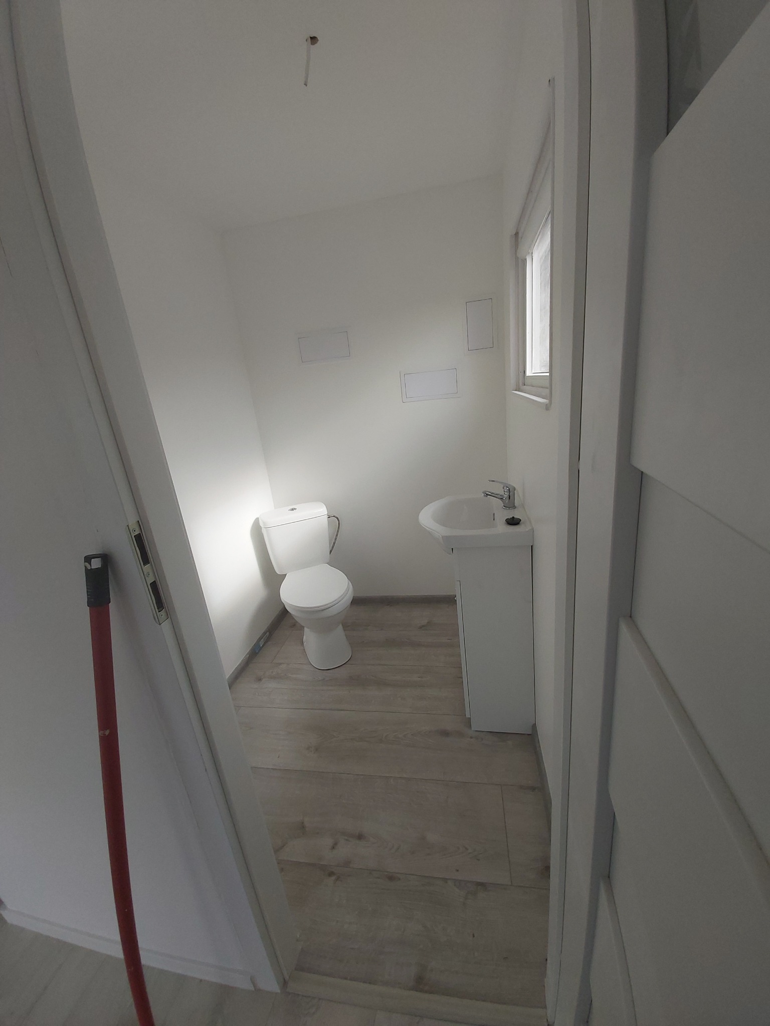 Jasna łazienka z toaletą, umywalką na szafce i drewnopodobną podłogą, widoczna przez otwarte drzwi, z czerwoną rurką opartą o framugę.