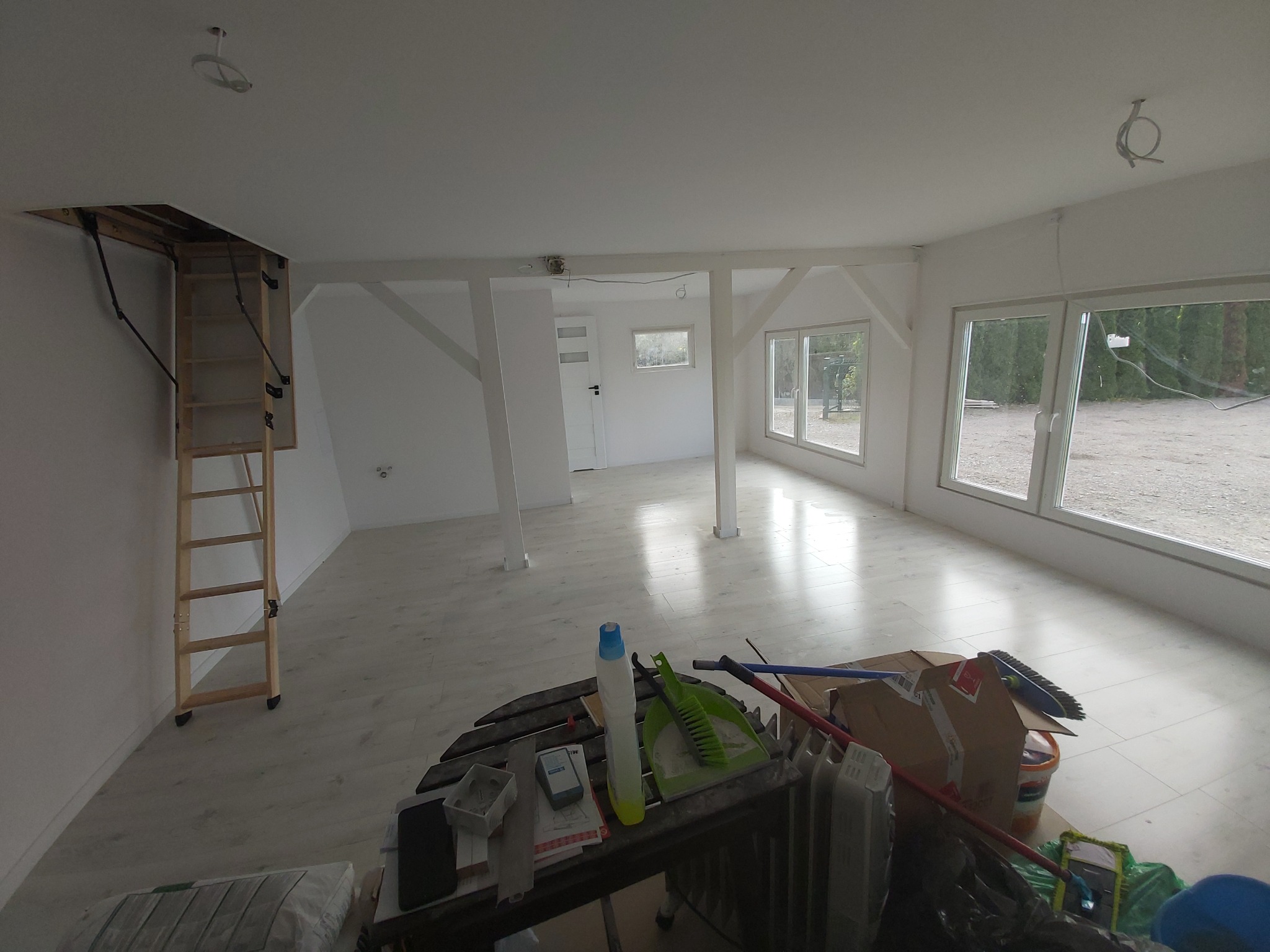 Surowe wnętrze z drewnianymi belkami, schodami strychowymi i dużymi oknami, częściowo umeblowane narzędziami i materiałami budowlanymi, ukazujące etap prac wykończeniowych.