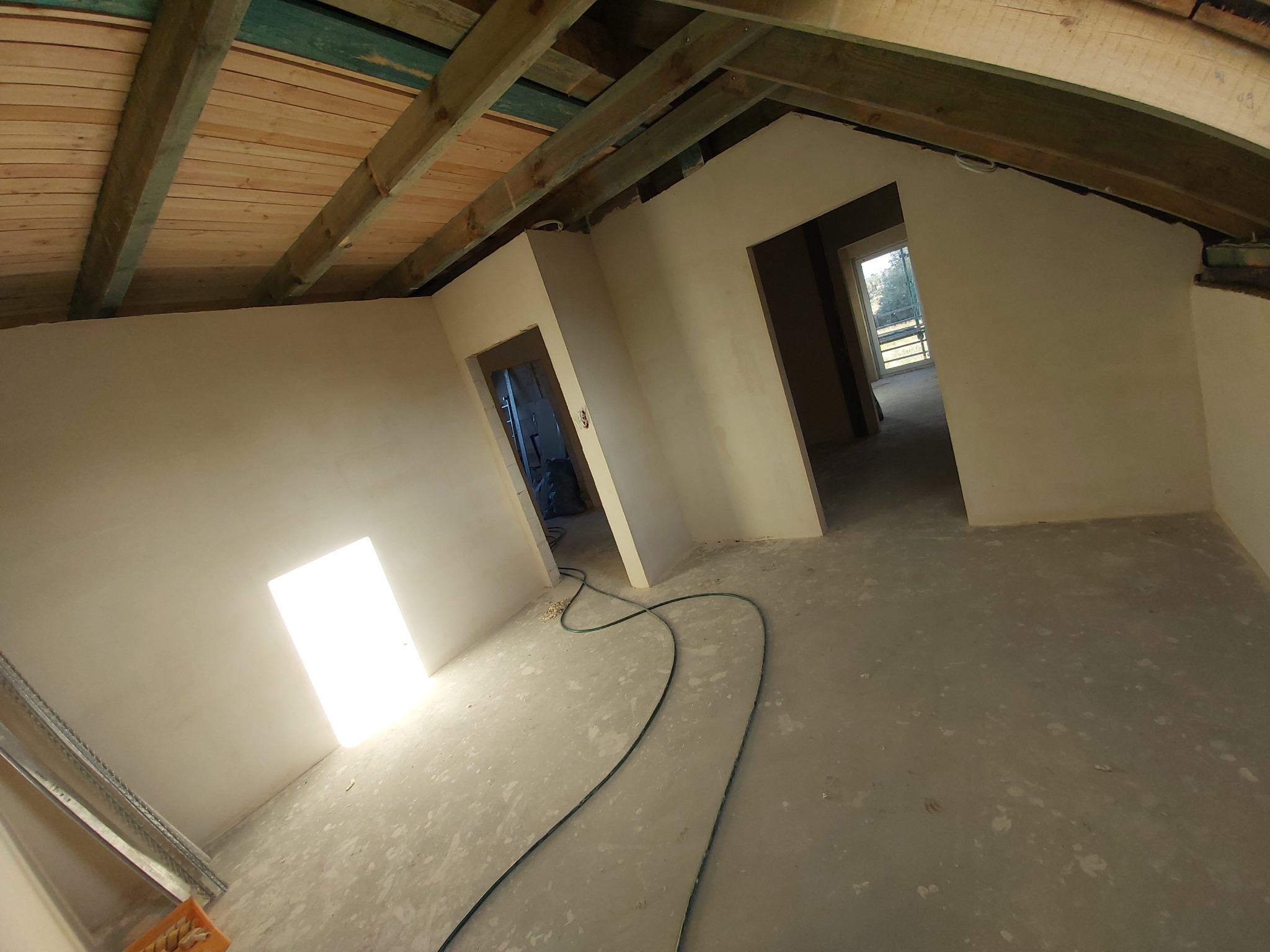 Surowe wnętrze poddasza w trakcie prac wykończeniowych, widoczne drewniane belki stropowe, otynkowane ściany z otworami drzwiowymi, szary betonowy podkład i przewód elektryczny na podłodze. W tle...