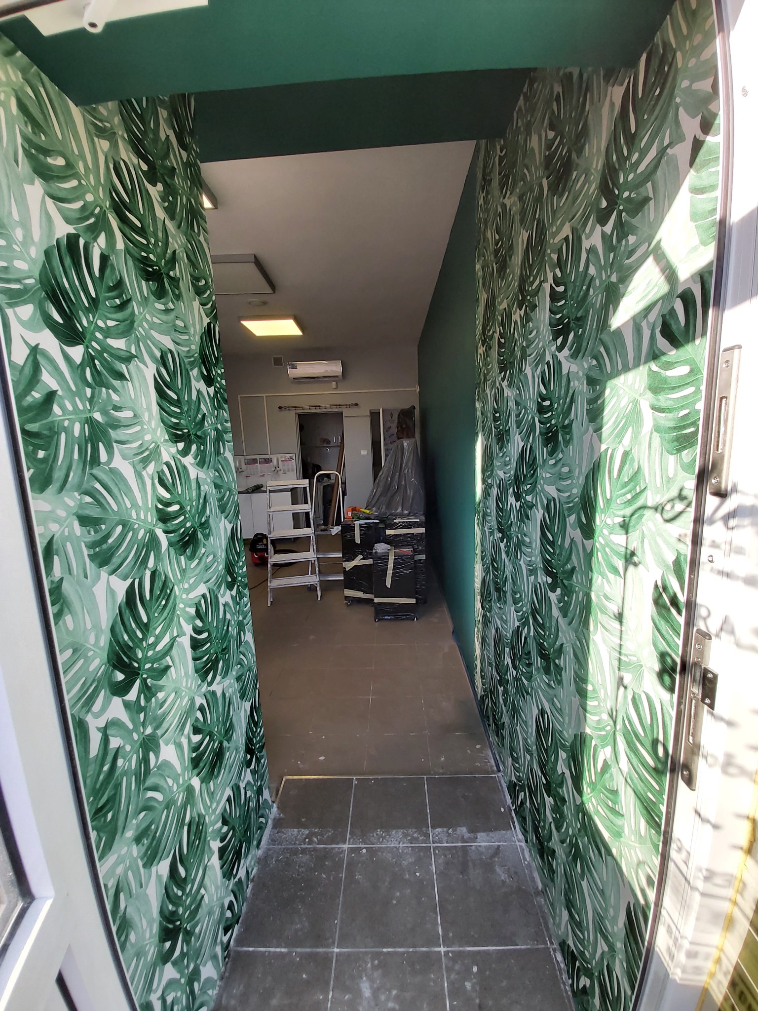 Korytarz z tapetą w motywy liści monstery, widoczne narzędzia i drabina w tle, świeżo pomalowany sufit na ciemnozielono.