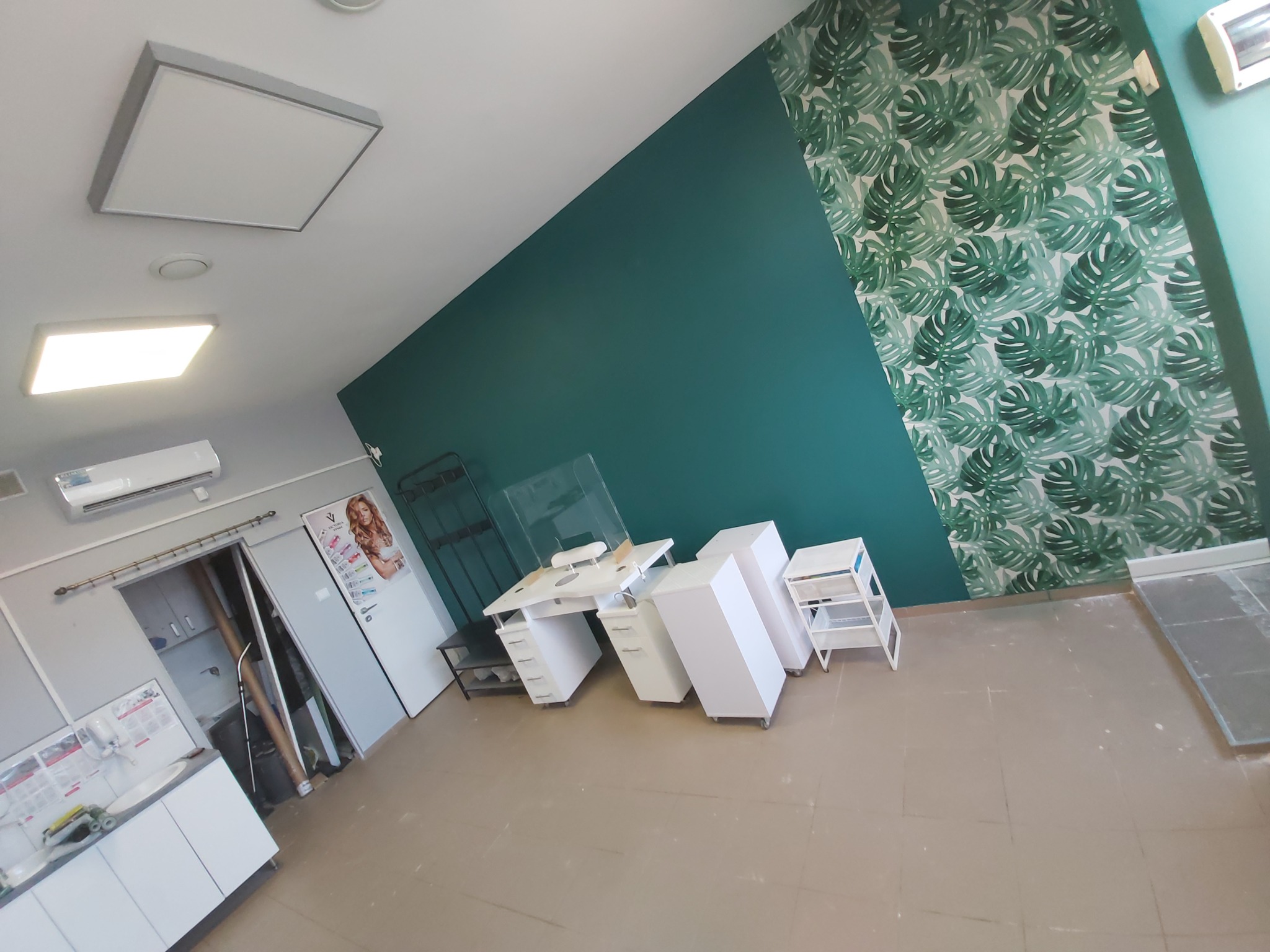 Wnętrze salonu kosmetycznego z białym biurkiem do manicure z pochłaniaczem pyłu, białymi szafkami na kółkach, ścianą w kolorze butelkowej zieleni i tapetą w liście monstery.