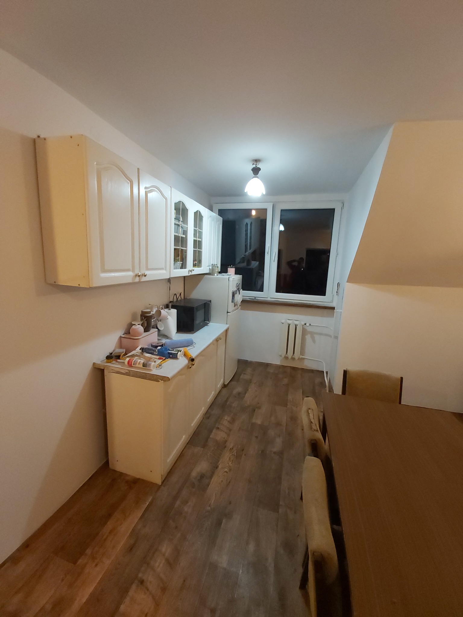 Kuchnia w trakcie remontu: białe szafki wiszące i stojące, narzędzia na blacie, widoczne ślady po malowaniu na ścianie, drewniana podłoga i fragment stołu w tle.