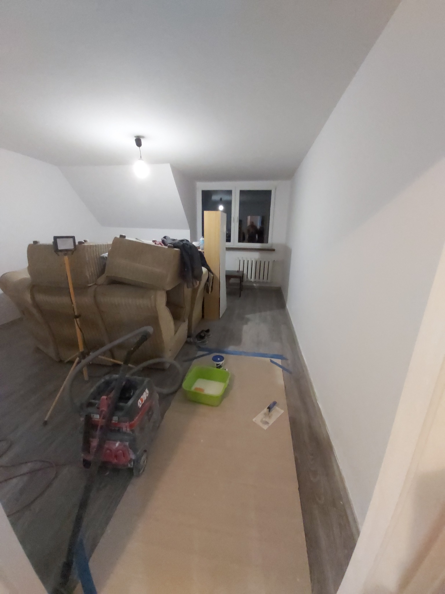 Pokój w trakcie remontu: świeżo pomalowane białe ściany, zabezpieczona podłoga, sprzęt malarski, sofa pod lampą budowlaną.