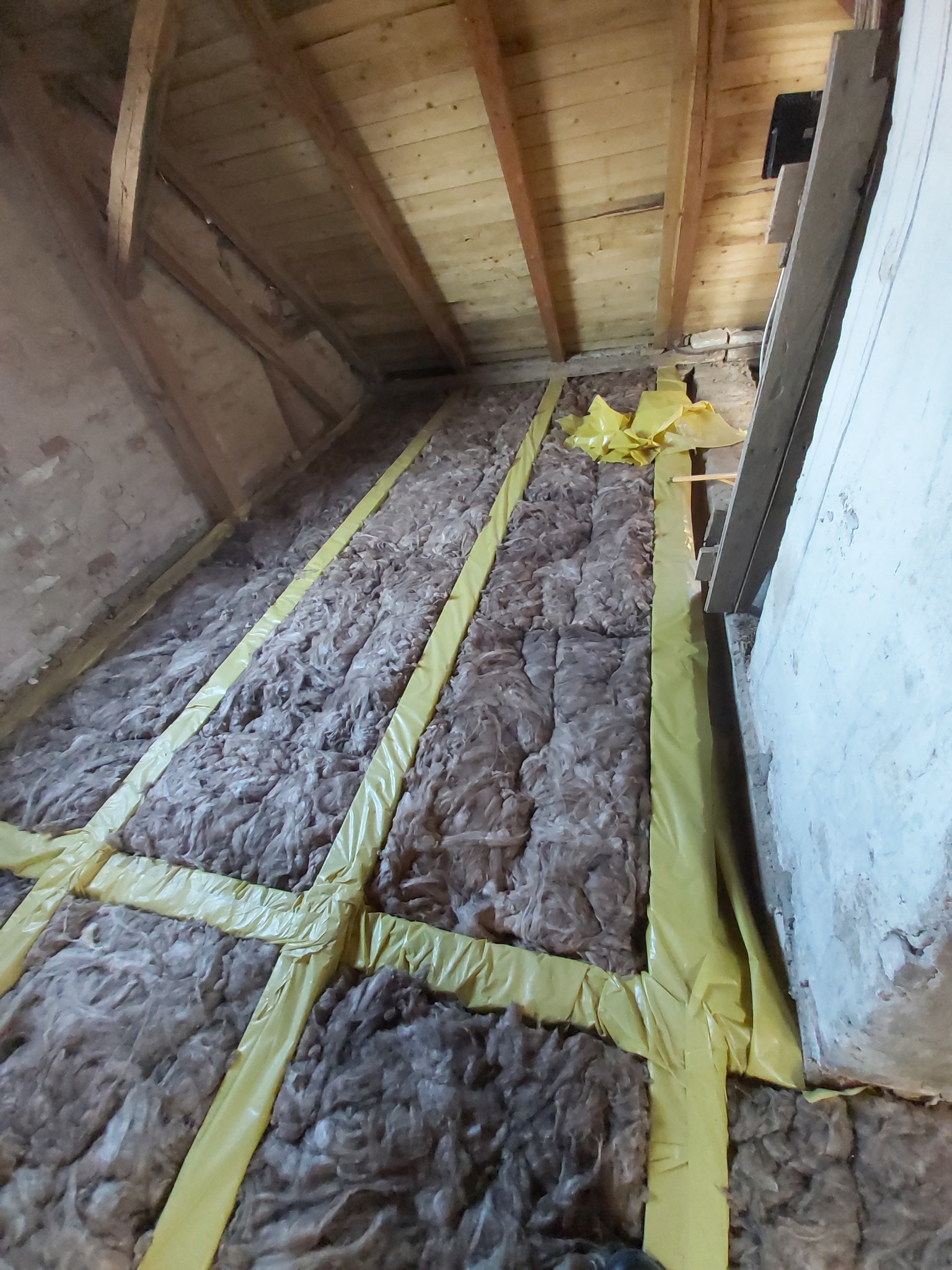 Ocieplenie poddasza wełną mineralną, zabezpieczone folią w kolorze żółtym, widoczne drewniane elementy konstrukcji dachu oraz fragment ściany.