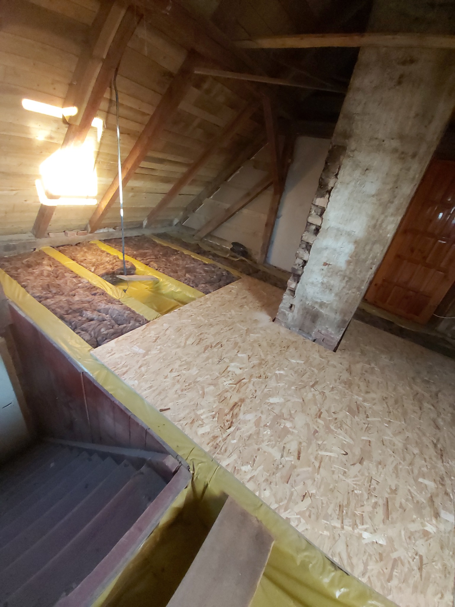 Poddasze w trakcie adaptacji: widoczna izolacja z wełny mineralnej zabezpieczona folią, fragment podłogi z płyt OSB oraz elementy konstrukcji dachu i komina.