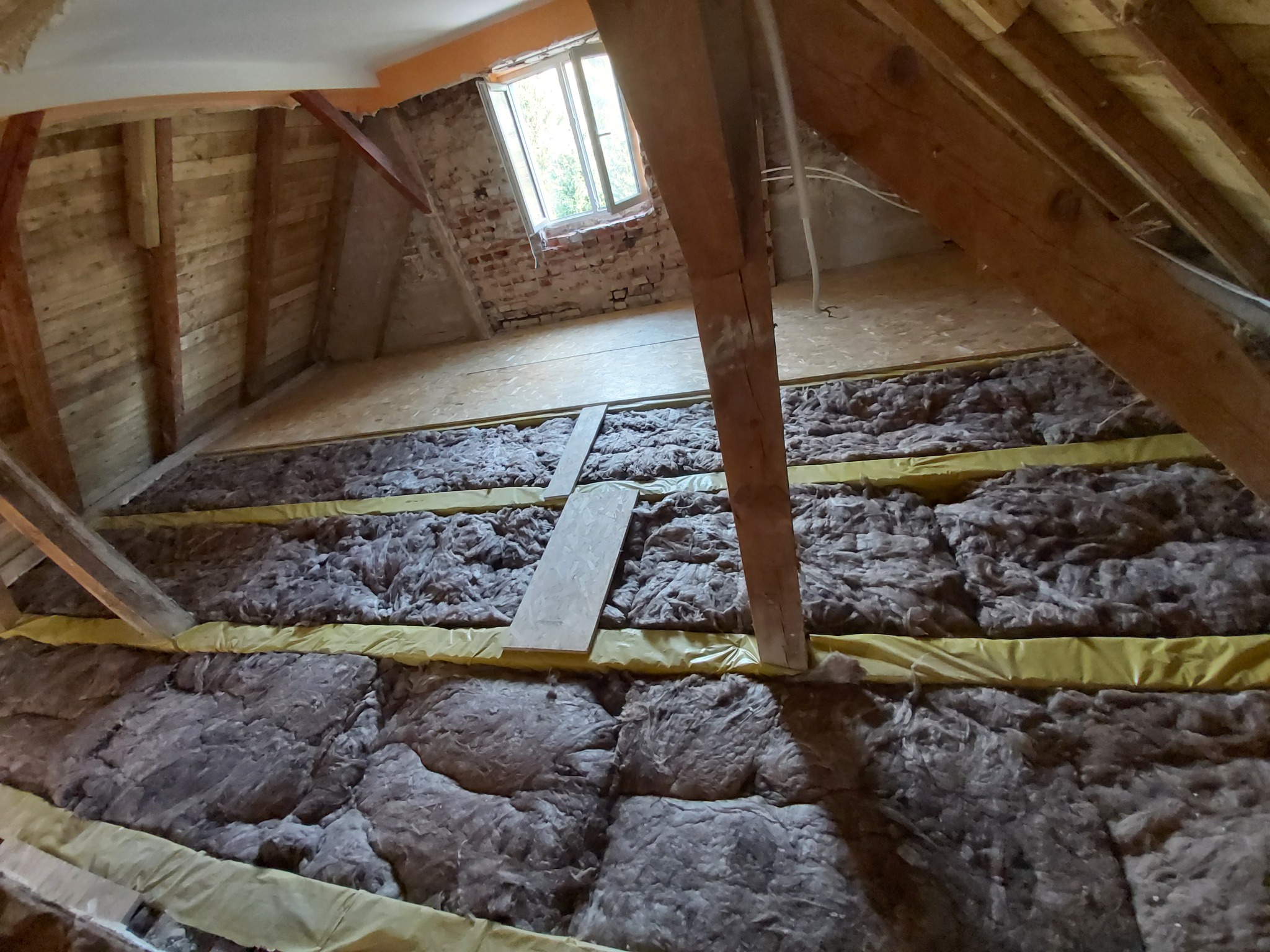 Izolacja poddasza wełną mineralną między krokwiami, widoczne folia paroizolacyjna i deski podłogowe, okno dachowe w tle.
