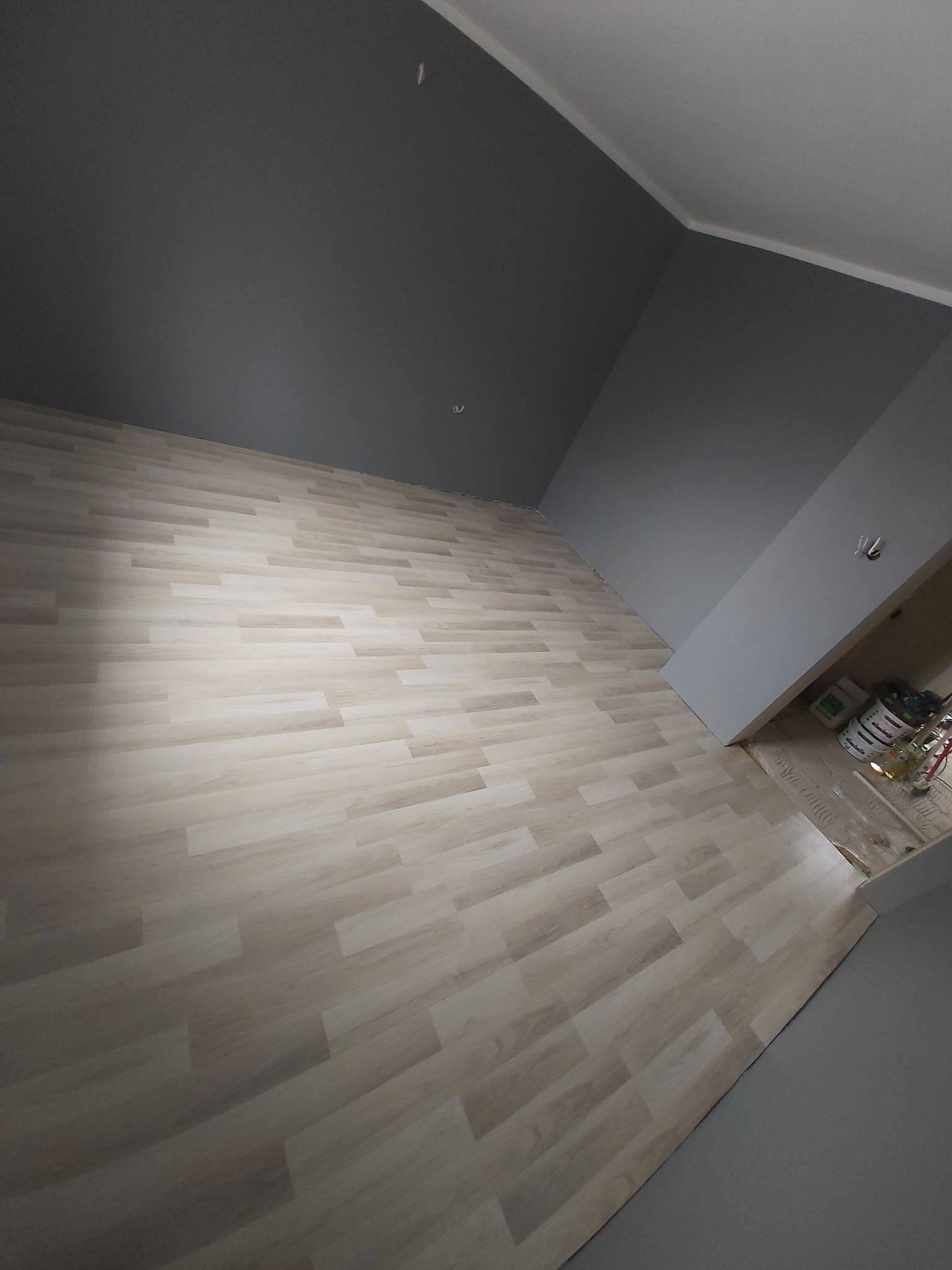 Świeżo pomalowane szare ściany i jasna podłoga panelowa w pustym pokoju, widoczne wiaderka z farbą w otwartych drzwiach.