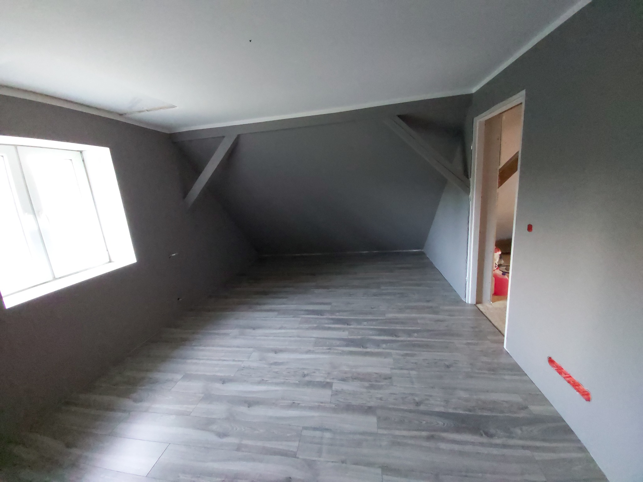 Szare, puste poddasze z drewnianą podłogą, skośnymi ścianami i widocznymi elementami konstrukcyjnymi, przygotowane do dalszego wykończenia.