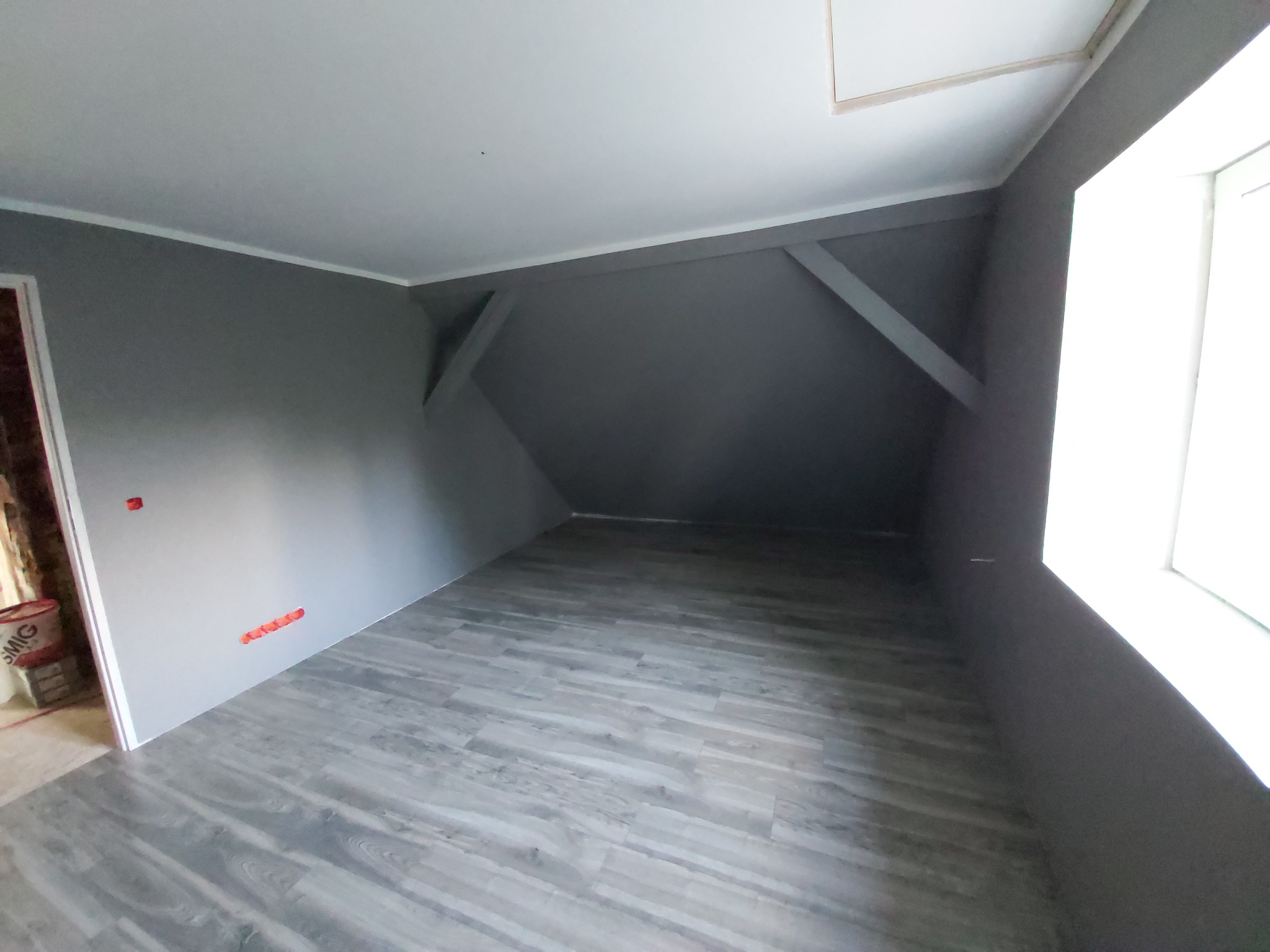 Wnętrze poddasza w Braniewie po malowaniu na szaro, z widoczną podłogą imitującą panele i fragmentem okna z prawej strony. Na ścianie widoczne punkty instalacji elektrycznej.