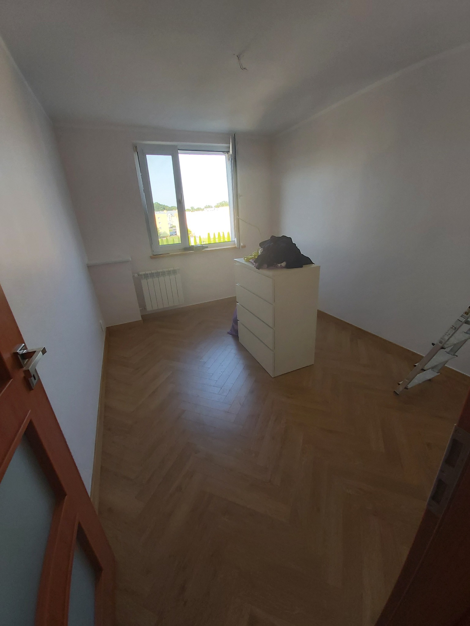 Pusty, jasny pokój z parkietem w jodełkę, białą komodą pod oknem i rozłożoną drabiną malarską przy ścianie.