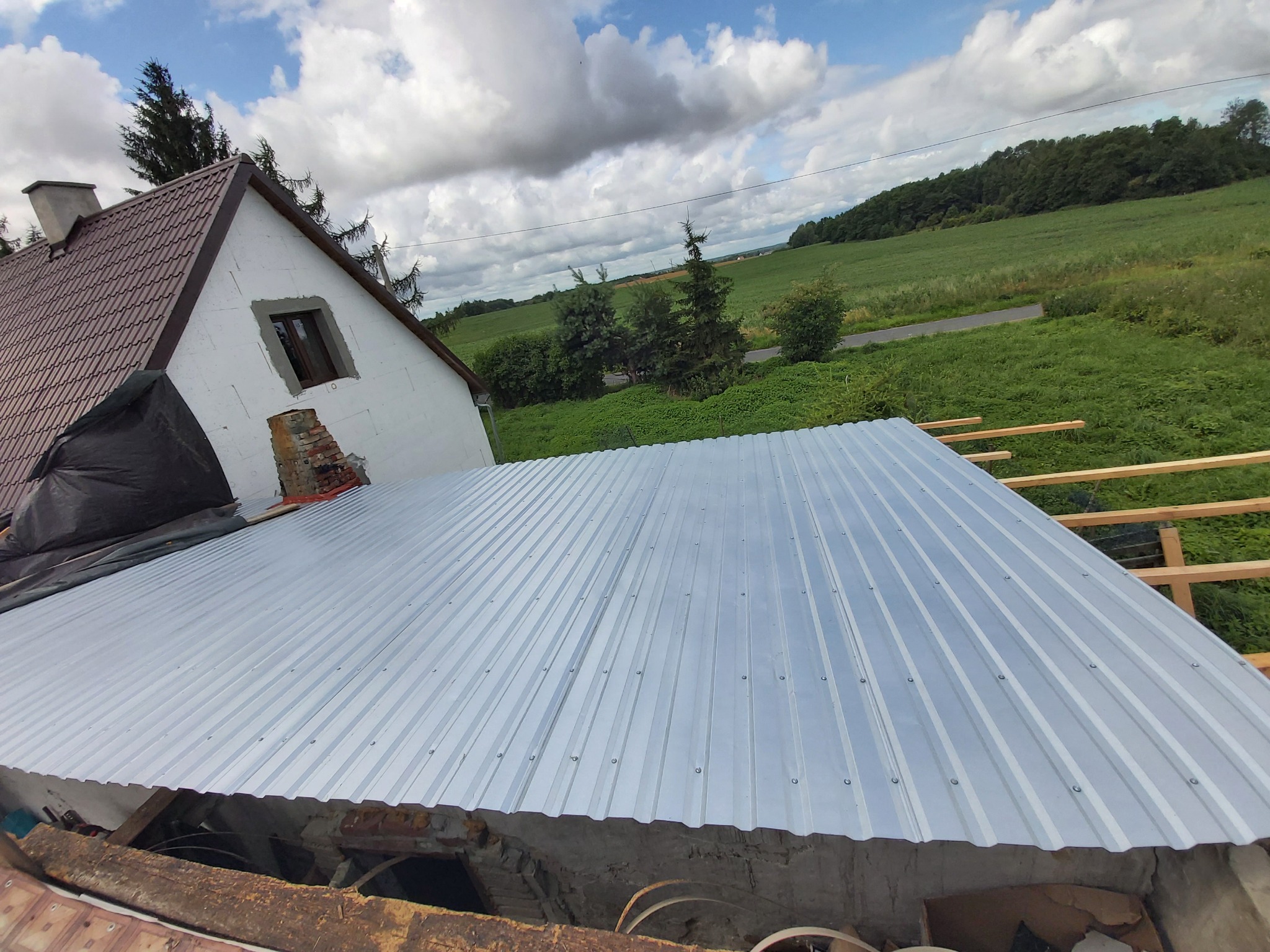 Montaż blachy trapezowej na dachu budynku gospodarczego z widoczną konstrukcją drewnianą i fragmentem murowanej ściany oraz otaczającym krajobrazem.