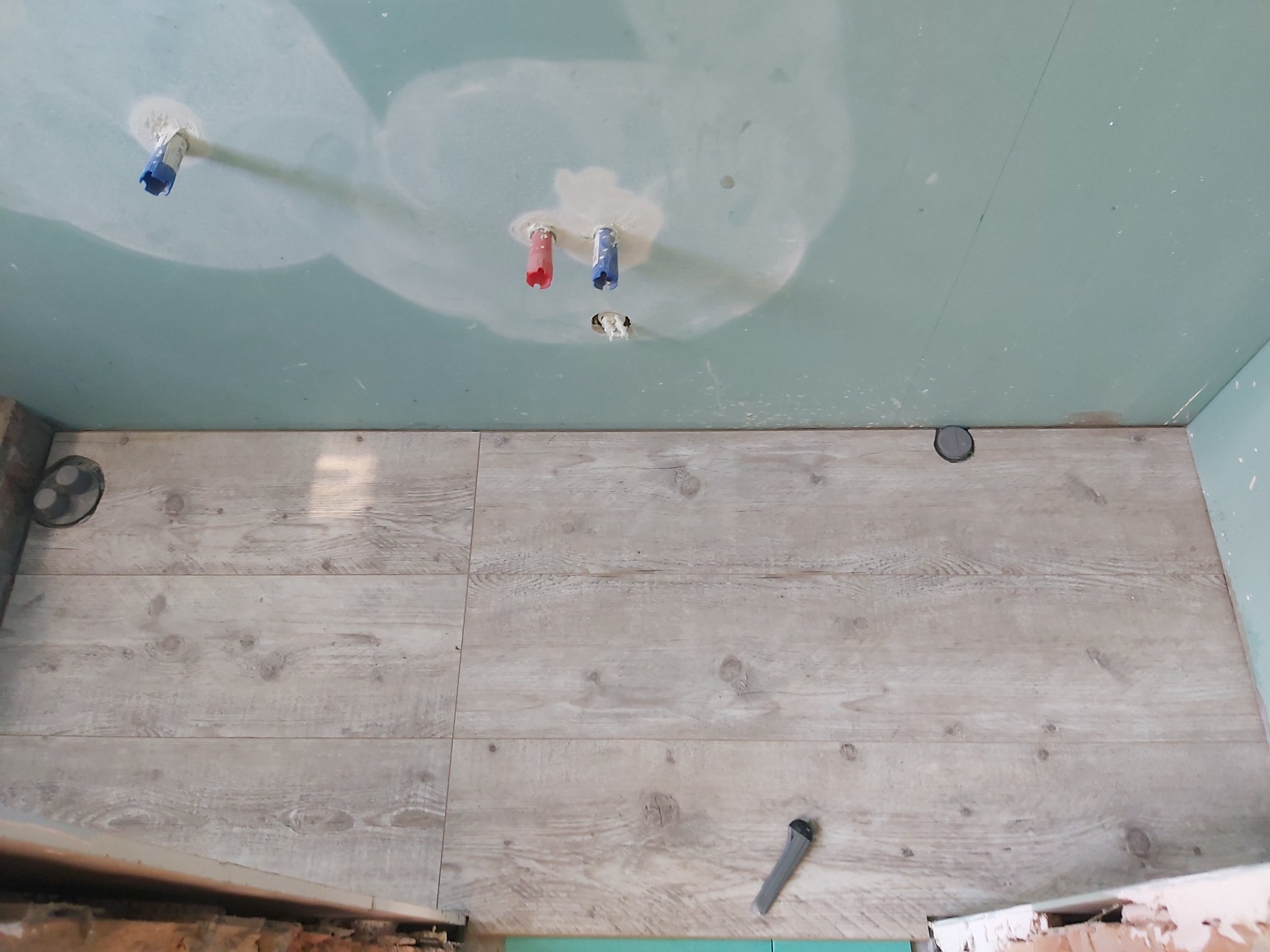 Niedokończona łazienka z położonymi panelami podłogowymi imitującymi drewno, widoczne wystające rury ze ściany i sufit z płyt kartonowo-gipsowych przed szpachlowaniem.