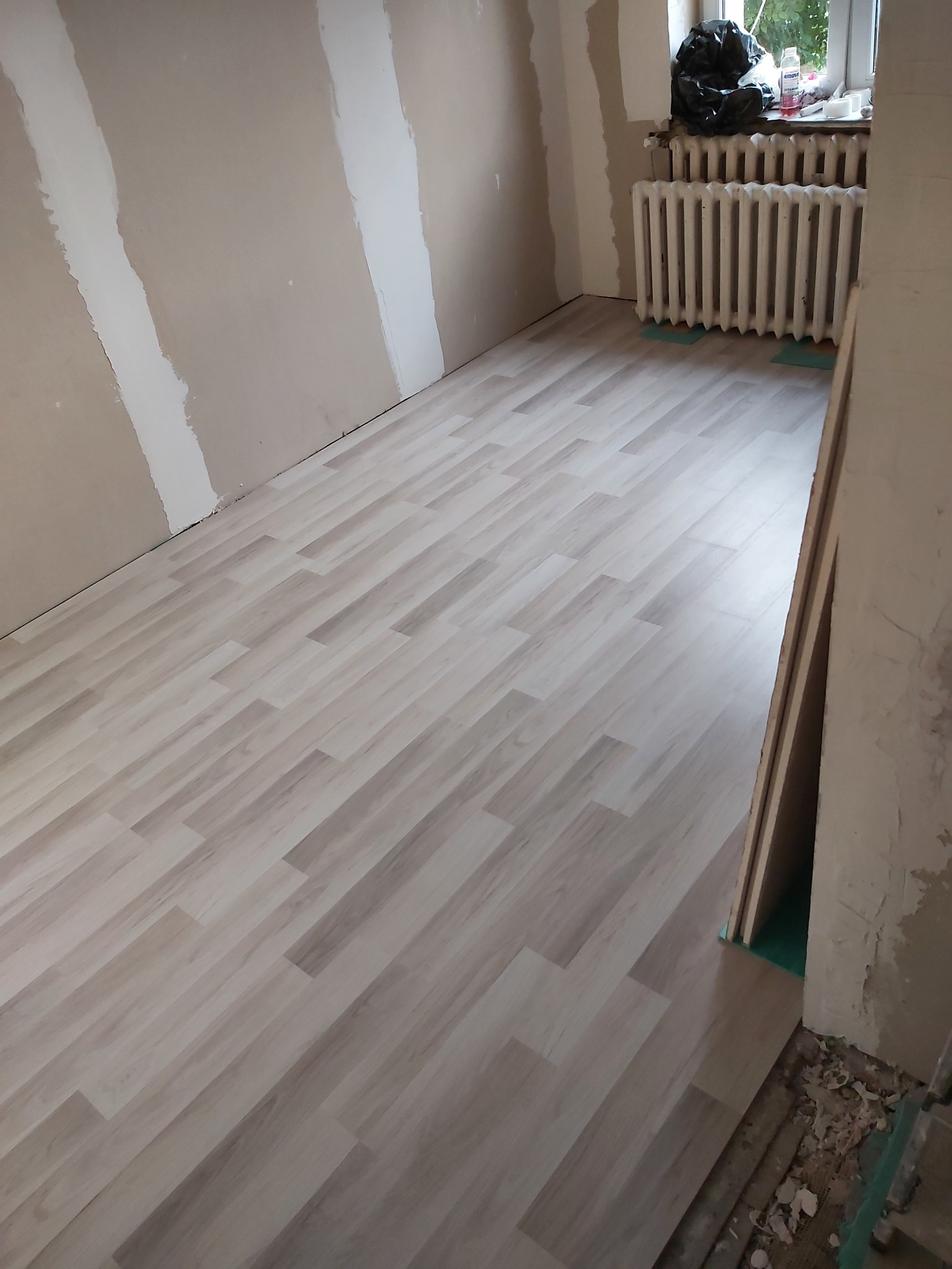 Wnętrze pokoju w trakcie remontu: jasna podłoga panelowa ułożona, ściany częściowo zagruntowane pod malowanie, widoczny grzejnik pod oknem i fragmenty gruzu przy futrynie.