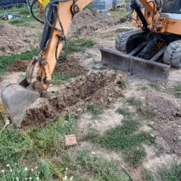 Pomarańczowa koparka w trakcie wykopu wąskiego rowu w ziemi, widoczne ślady pracy i stosy ziemi, w tle zaparkowany samochód osobowy i palety z kostką brukową.
