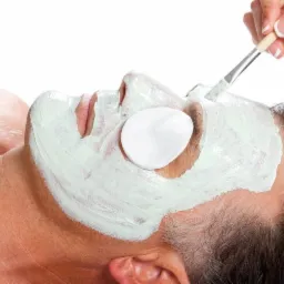 Mężczyzna z nałożoną zieloną maską na twarzy, na jednym oku wacik, kosmetyczka nakłada maskę pędzlem.