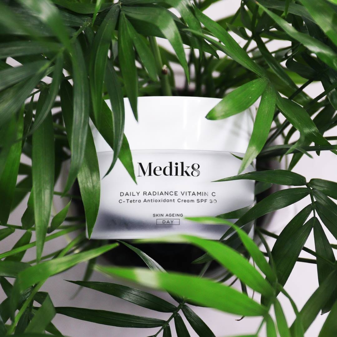 Biały słoiczek kremu Medik8 Daily Radiance Vitamin C SPF 30 otoczony zielonymi liśćmi rośliny doniczkowej, z widocznym logo i nazwą produktu, sugerujący pielęgnację skóry.