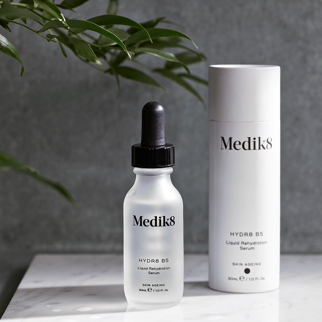 Dwa produkty Medik8 HYDR8 B5 Liquid Rehydration Serum: buteleczka z pipetą i tuba, na marmurowym blacie, z zieloną gałązką w tle.