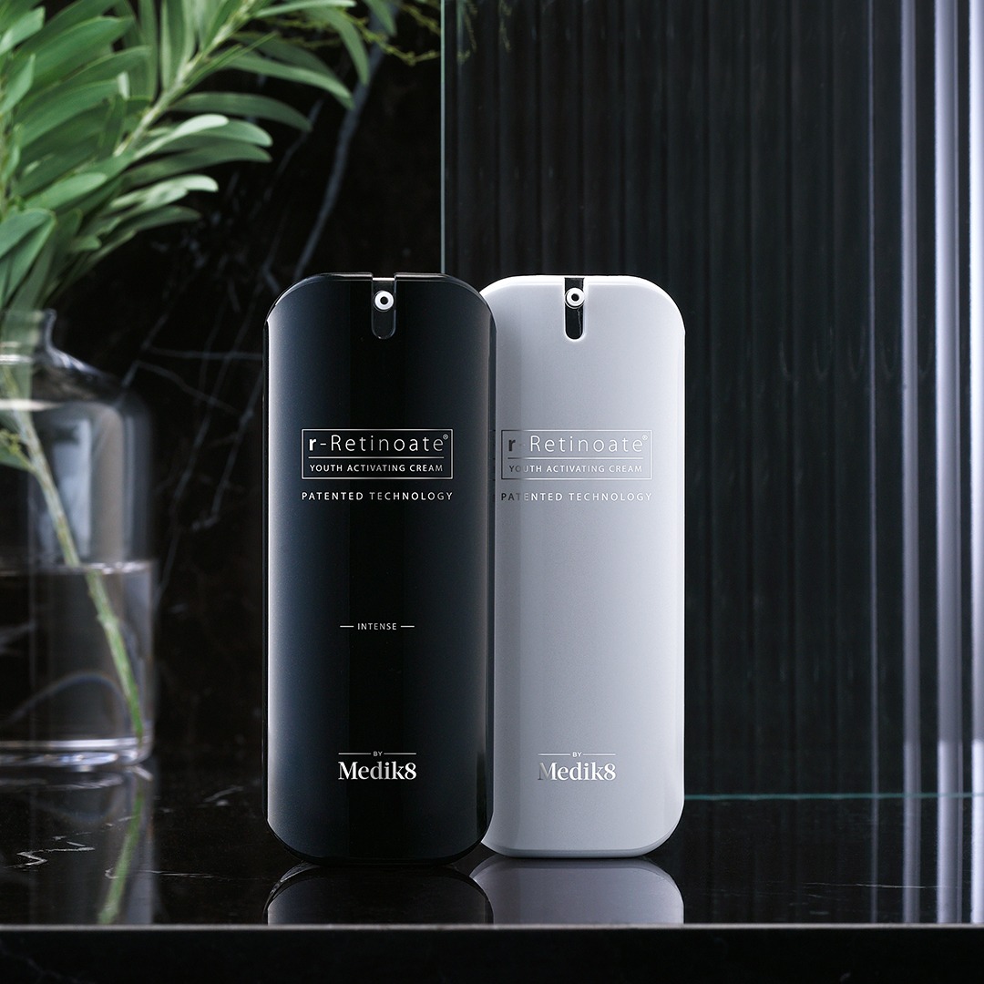 Dwie eleganckie buteleczki kremu r-Retinoate Youth Activating Cream marki Medik8, jedna czarna, druga biała, ustawione na błyszczącej, marmurkowej powierzchni, z rośliną w tle.