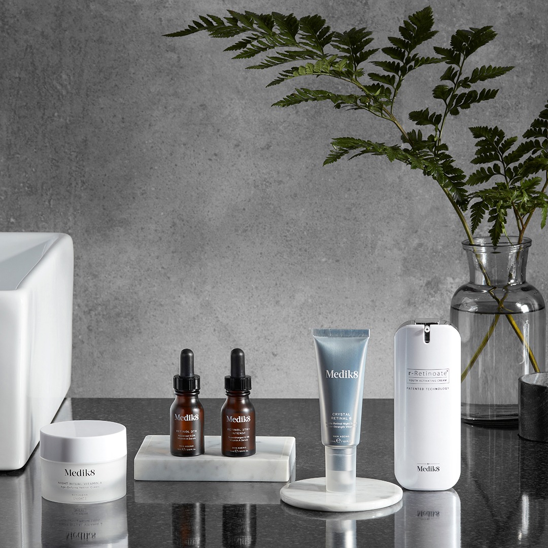 Zestaw kosmetyków marki Medik8 na marmurowym blacie: krem, serum z pipetą, tubka i dozownik, uzupełniony o wazon z paprocią na tle szarej ściany.