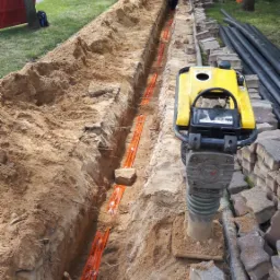 Głęboki wykop w ziemi z pomarańczową taśmą ostrzegawczą, obok ubijarka wibracyjna i ułożone krawężniki, w tle pracownik przy wykopie i koparka.