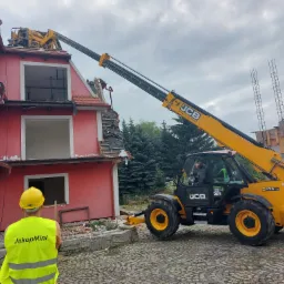 Żółty podnośnik teleskopowy JCB demontuje fragment dachu czerwonego, dwupiętrowego domu. Pracownik w żółtym kasku i kamizelce obserwuje postęp prac. Widoczne uszkodzone fragmenty dachu i ściany...