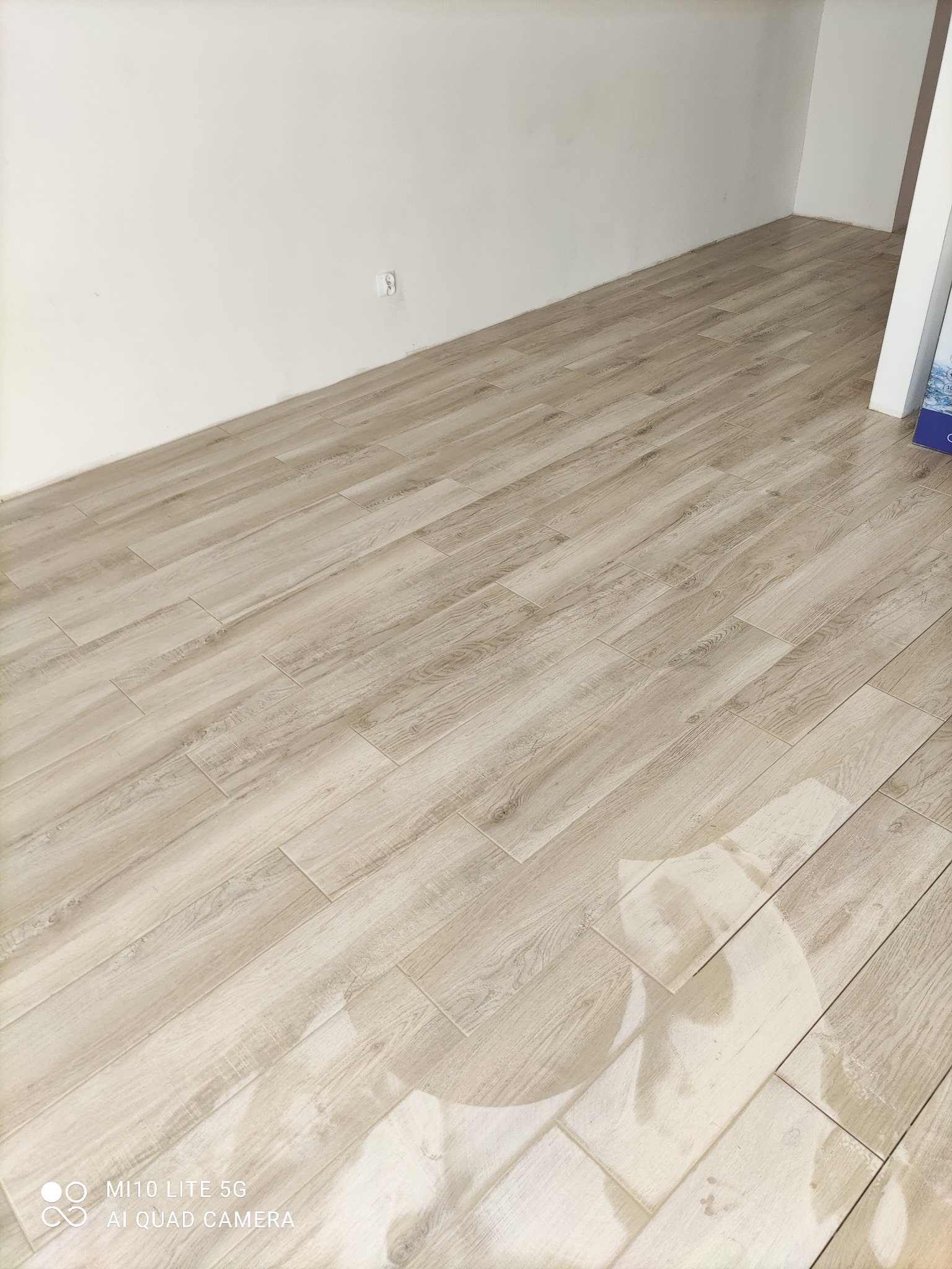 Ułożone panele podłogowe imitujące jasne drewno w pustym pomieszczeniu z białymi ścianami, widoczny fragment ściany z gniazdkiem elektrycznym.