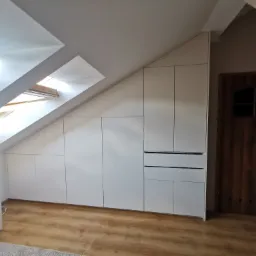 Biała zabudowa meblowa pod skosem na poddaszu, z widocznymi oknami dachowymi i drewnianymi drzwiami po prawej stronie.