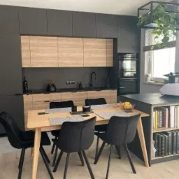Nowoczesna kuchnia z szarymi szafkami górnymi i dolnymi z drewnopodobnym frontem, czarnym blatem i zlewem, uzupełniona o stół z czarnymi krzesłami i regał z książkami.