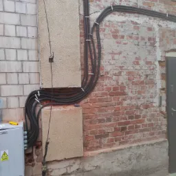 Zewnętrzna instalacja elektryczna: czarne rury karbowane przymocowane do ściany z cegły i bloczków, z puszką elektryczną i żółtym miernikiem w pobliżu.