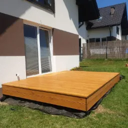 Nowoczesny taras z desek w kolorze naturalnego drewna, przylegający do budynku z dużym oknem tarasowym, widoczna czarna mata pod spodem.