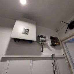 Biały falownik FOX-ESS z niebieskimi złączami, skrzynka bezpiecznikowa z otwartą obudową i skrzynka KENO z żółtą naklejką ostrzegawczą zamontowane na ścianie wewnątrz budynku.